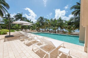 Un espace aménagé au bord de la piscine, avec des chaises longues et des parasols, entouré de palmiers et surplombé d’un ciel dégagé.