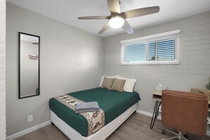 Un dormitorio con una cama individual, ventilador de techo, ventana con persianas, escritorio con silla y espejo en la pared.