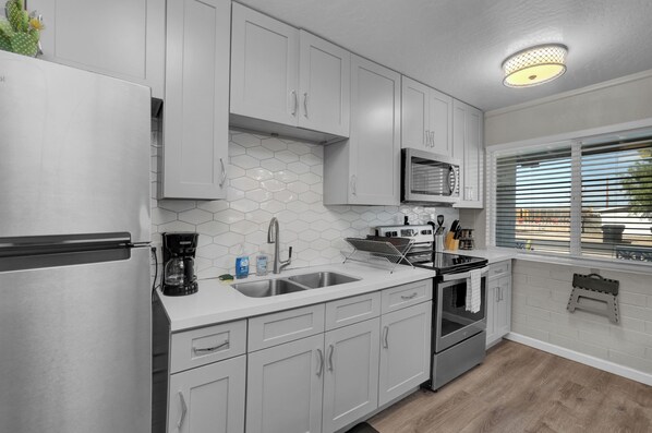 A modern kitchen with white cabinets, a stainless steel refrigerator, a microwave, and a window with blinds.