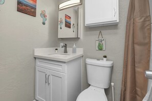 Ein Badezimmer mit einem weißen Waschbecken, einer Toilette und einem Spiegel über dem Waschbecken. An den Wänden befinden sich Dekorationen, darunter ein Kaktus und ein Gemälde.