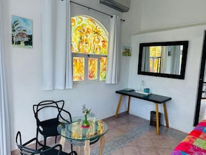 Una habitación con una mesa de vidrio, dos sillas, un escritorio, un espejo y una ventana con cortinas.