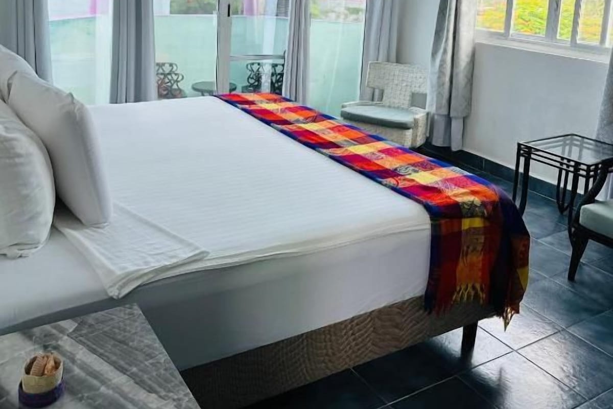 Habitación de hotel con una cama grande, ventilador de techo, ventana con cortinas y vistas al exterior.