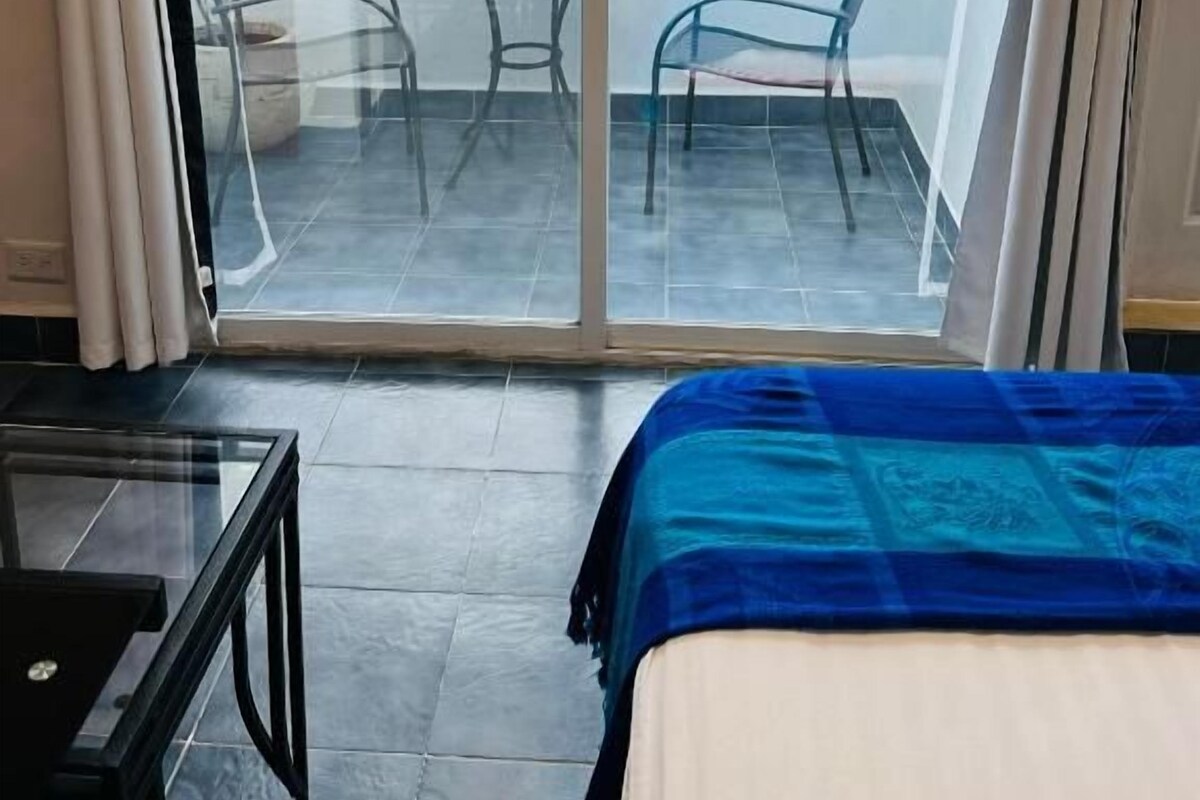 A room with a bed, a glass door leading to an outdoor area with a table and chairs, and a view of trees and flowers.