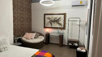 A hotel room with a bed, a chair, a small table, a wall-mounted air conditioner, and a framed abstract painting.
