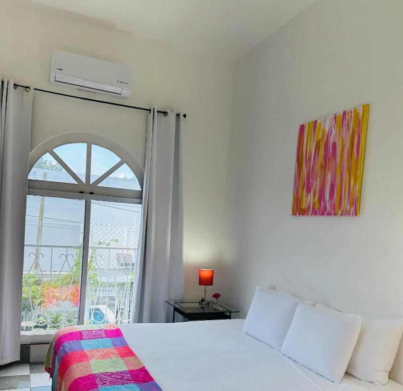 Hotelkamer met een bed, een raam met gordijnen, een airconditioner, een nachtkastje met een lamp en een kleurrijk abstract schilderij aan de muur.