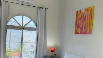 Hotelkamer met een bed, een raam met gordijnen, een airconditioner, een nachtkastje met een lamp en een kleurrijk abstract schilderij aan de muur.