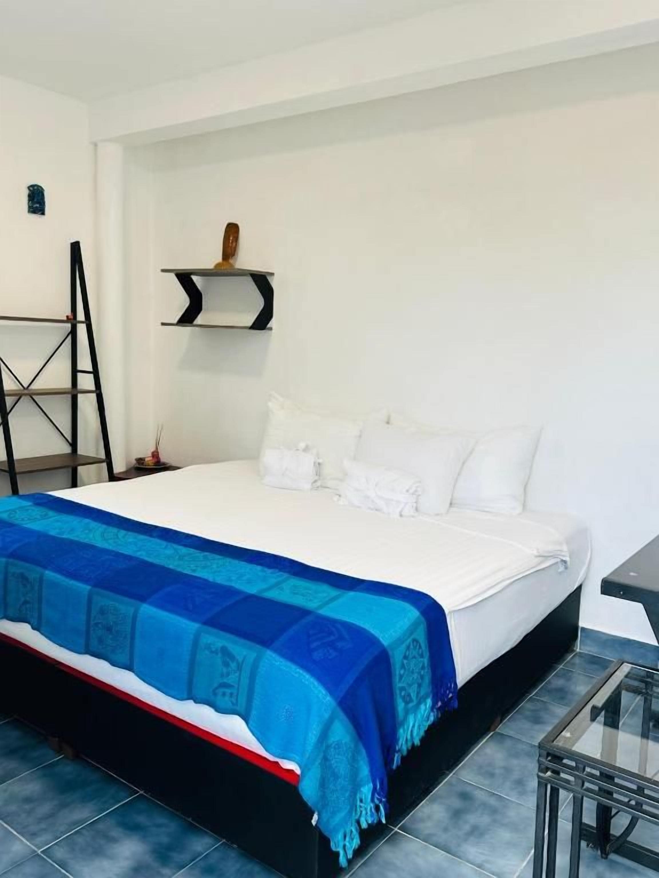 A bedroom with a bed, a blue blanket, a ladder, a shelf with decorative items, and a glass coffee table.