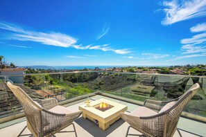 A balcony with wicker chairs, a fire pit, and a glass railing overlooking a coastal landscape. - Soledad Sky House | Pool + Sweeping Ocean Views (San Diego)