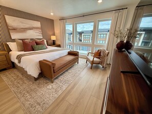 A modern bedroom with a large bed, a wooden headboard, a bedside table, a nightstand, a sofa, a chair, a window with a view of buildings, and a wooden floor. - 3-Bedroom Townhouse with Mountain and Lake Views. Sleep 9 (Heber City)