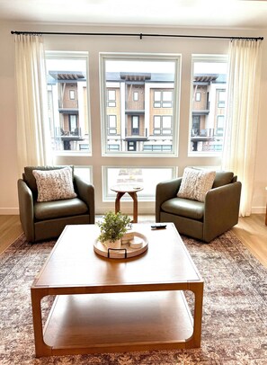 A living room with two leather sofas, a wooden coffee table, a small round table, and a view of a residential building through large windows. - 3-Bedroom Townhouse with Mountain and Lake Views. Sleep 9 (Heber City)