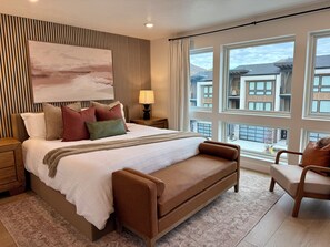 A modern bedroom with a large bed, a nightstand, a lamp, and a view of buildings through large windows. - 3-Bedroom Townhouse with Mountain and Lake Views. Sleep 9 (Heber City)