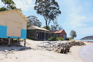 Un bâtiment situé sur la côte, avec un toit en métal ondulé, une structure en bois et une porte bleue, ainsi qu’une plage de sable avec des rochers et un ciel dégagé.