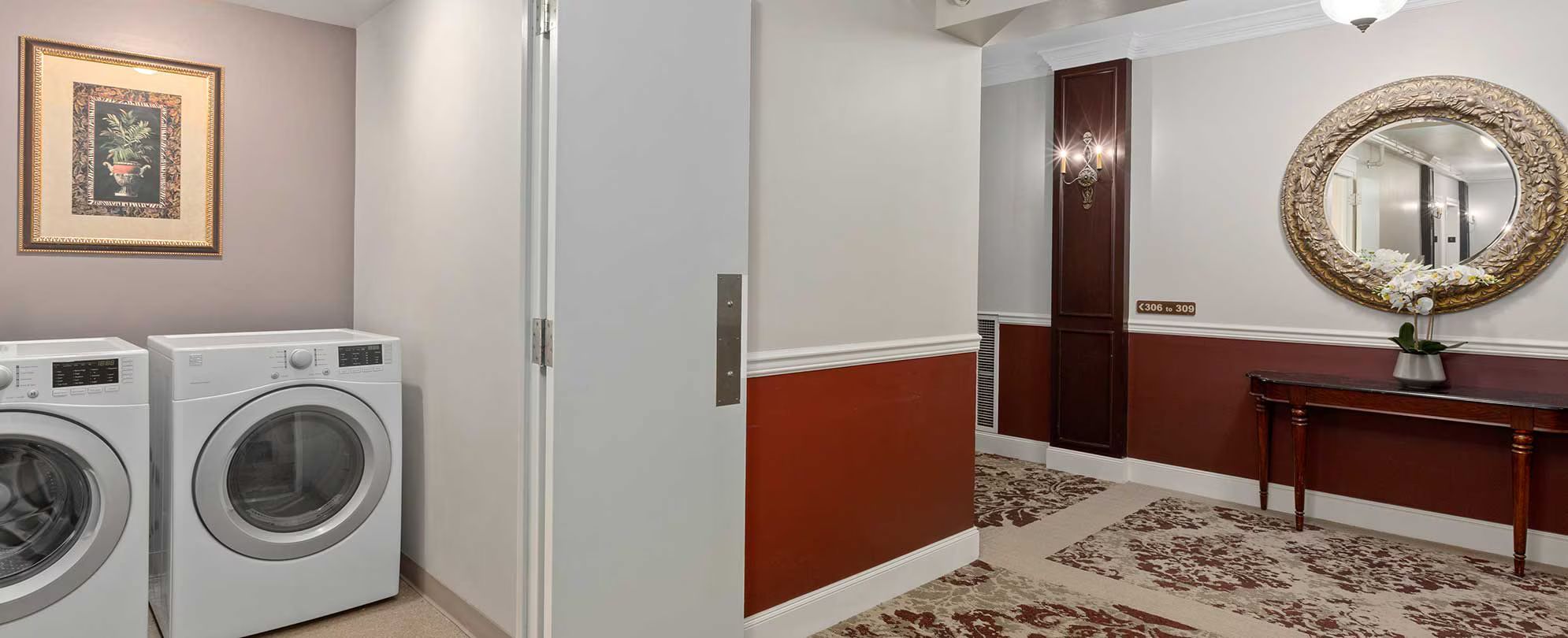 A hotel room with a washing machine, a mirror with an ornate frame, and a door with a number sign.