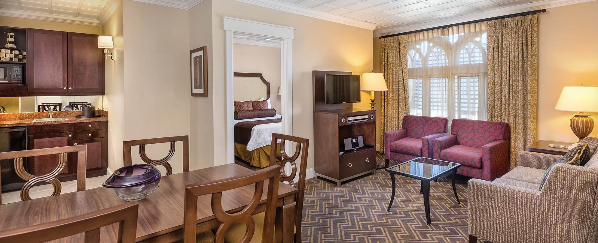 A hotel room with a dining area, a sofa set, a television, and a large window with curtains.