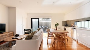 A modern living space with a sofa, dining table, chairs, a television, and a kitchen area. - Cuthbert Place Broadmeadows (Broadmeadows)