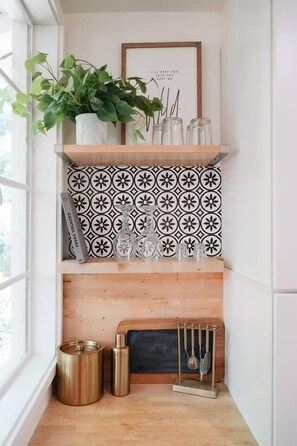 A corner with a wooden shelf holding a potted plant, a framed quote, and a patterned tile backsplash. Below, a fireplace with a metal stand and a small trash can are visible. - Nature Lovers Casita! King Bed! Big Backyard (Fresno)