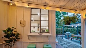 A covered outdoor seating area with a window overlooking a garden, a grill, and a bench.