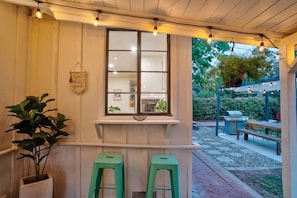A covered outdoor seating area with a window overlooking a garden, a grill, and a bench.