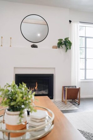 A living room with a fireplace, a round mirror on the wall, a potted plant, and a window with curtains. - Nature Lovers Casita! King Bed! Big Backyard (Fresno)