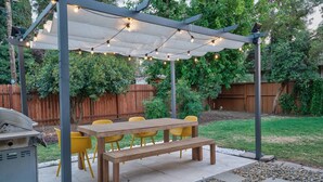 A covered outdoor patio with a wooden table and yellow chairs, surrounded by a garden and a wooden fence.
