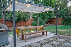 A covered outdoor patio with a wooden table and yellow chairs, surrounded by a garden and a wooden fence.