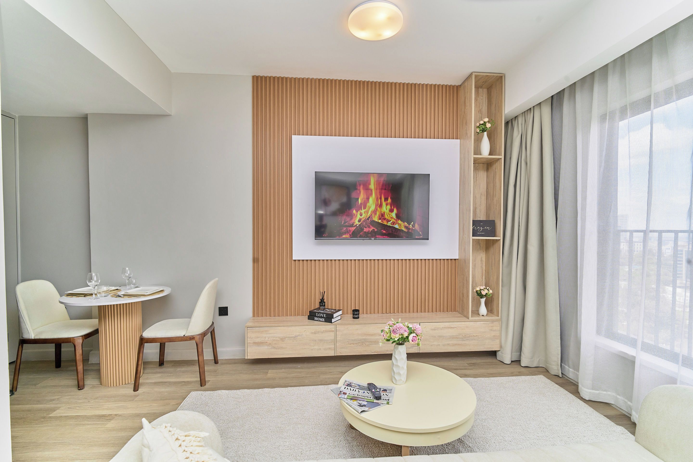 A modern living room with a flat-screen TV mounted on a wooden panel wall, a round coffee table with a vase and remote control, a dining area with a table set for two, and a large window with sheer curtains.
