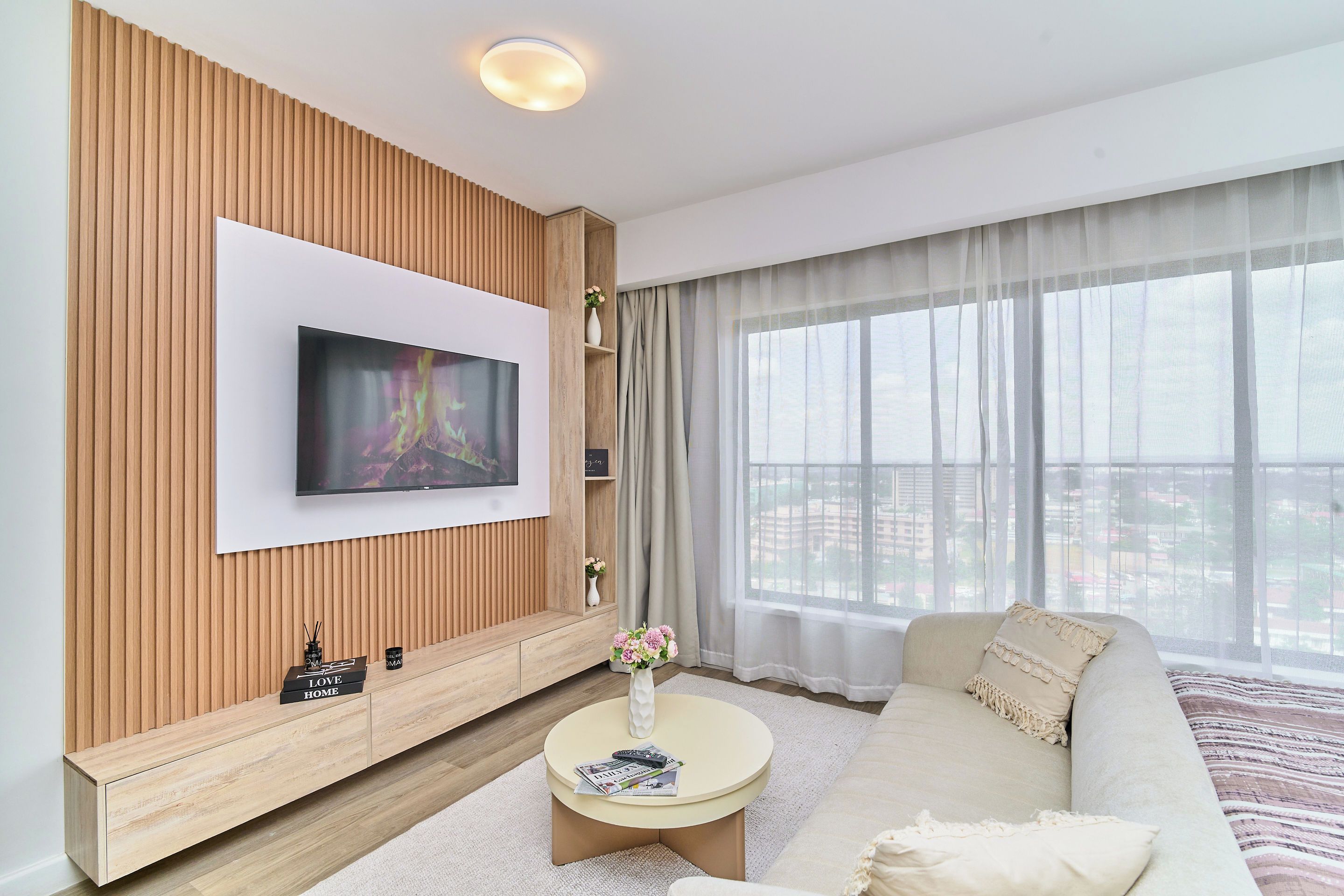 A modern living room with a flat-screen TV mounted on a wooden panel wall, a beige sofa, a round coffee table, and a large window with sheer curtains.