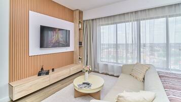 A modern living room with a flat-screen TV mounted on a wooden panel wall, a beige sofa, a round coffee table, and a large window with sheer curtains.