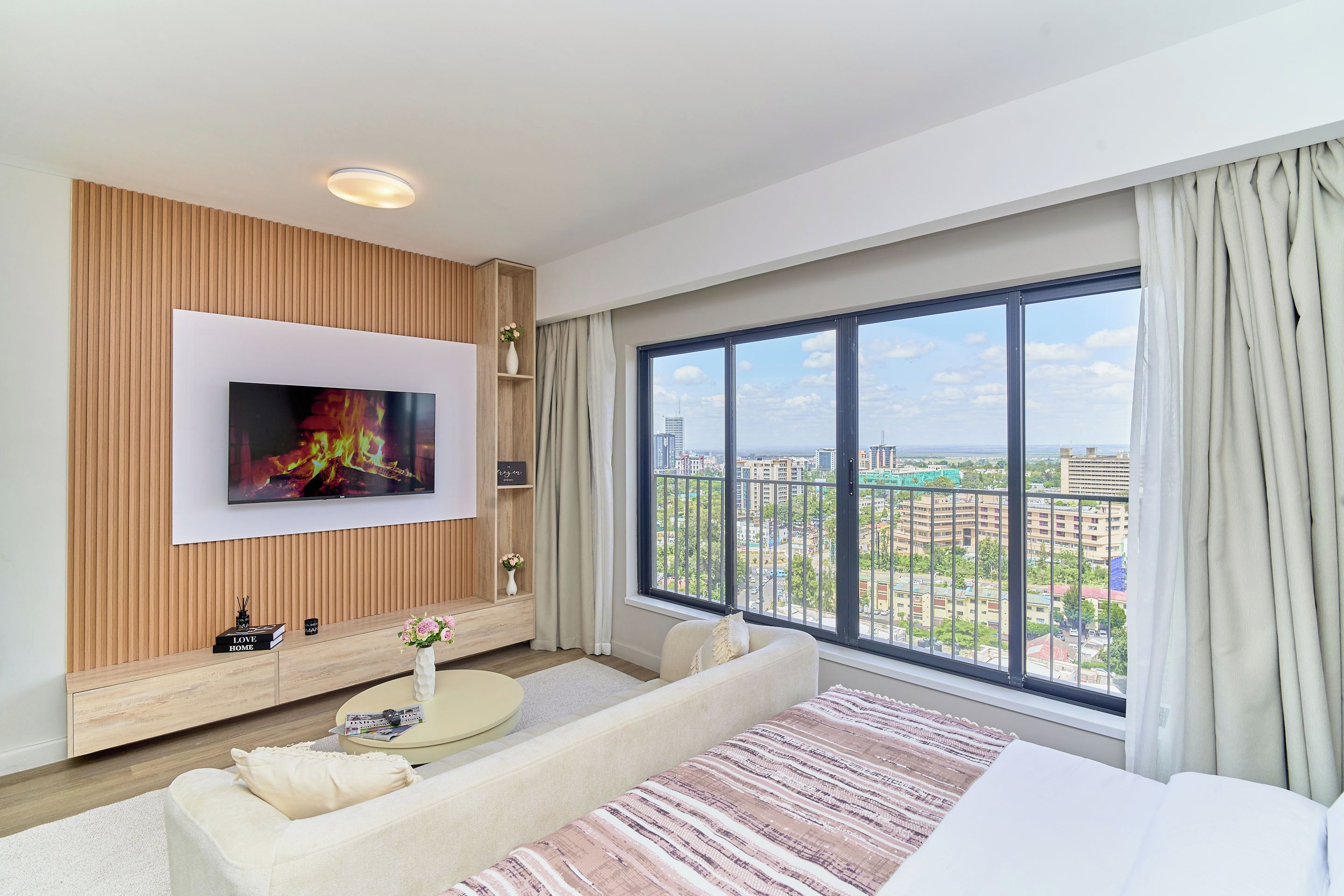 A modern living room with a flat-screen TV mounted on a wooden panel wall, a sofa, a coffee table, and a large window with a city view.