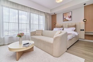 Une chambre d’hôtel moderne avec un grand lit, un canapé, une table basse ronde et une vue sur la ville à travers de fines rideaux.
