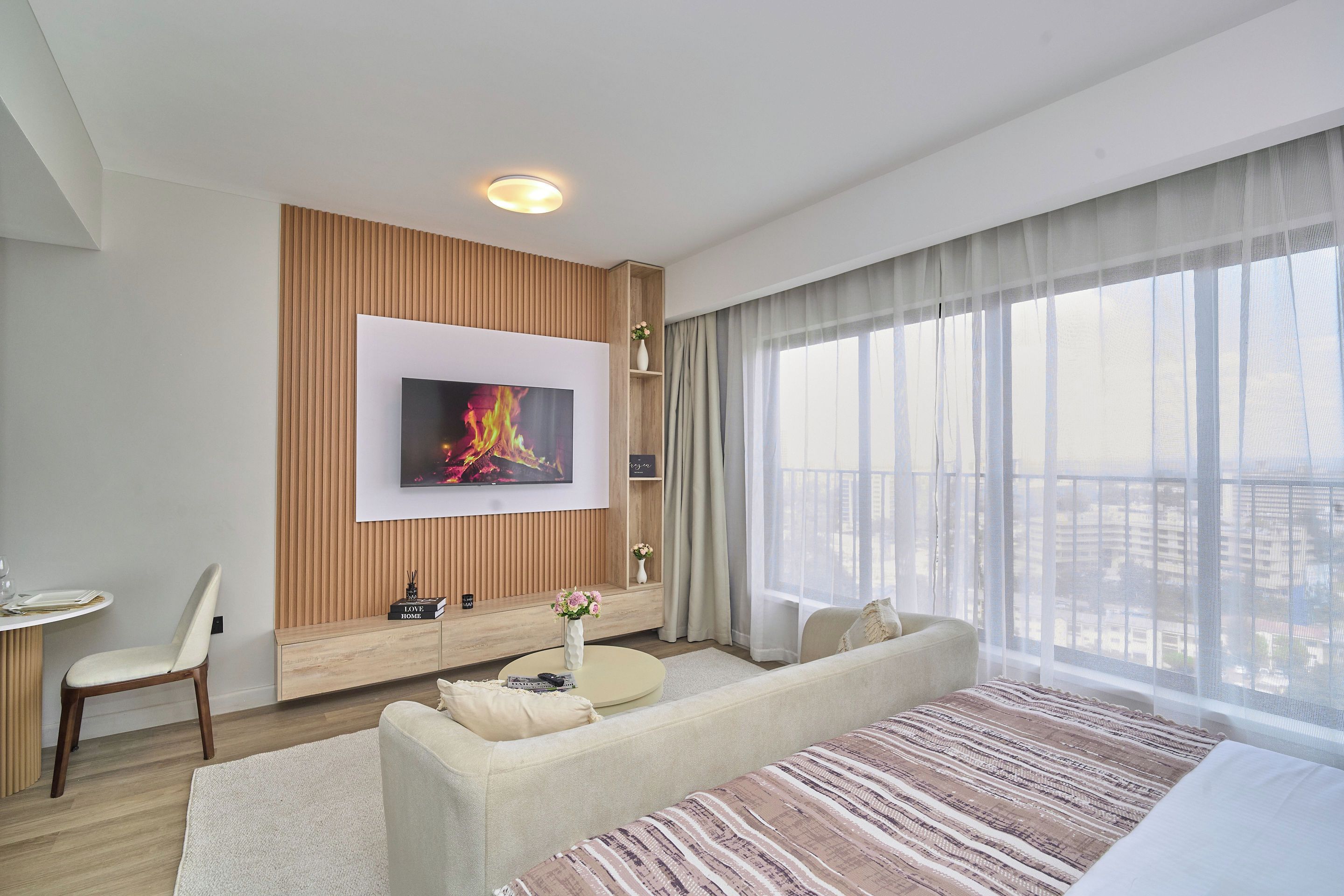 A modern hotel room with a flat-screen TV mounted on a wooden panel wall, a sofa, a small table with a vase of flowers, and a large window with sheer curtains.