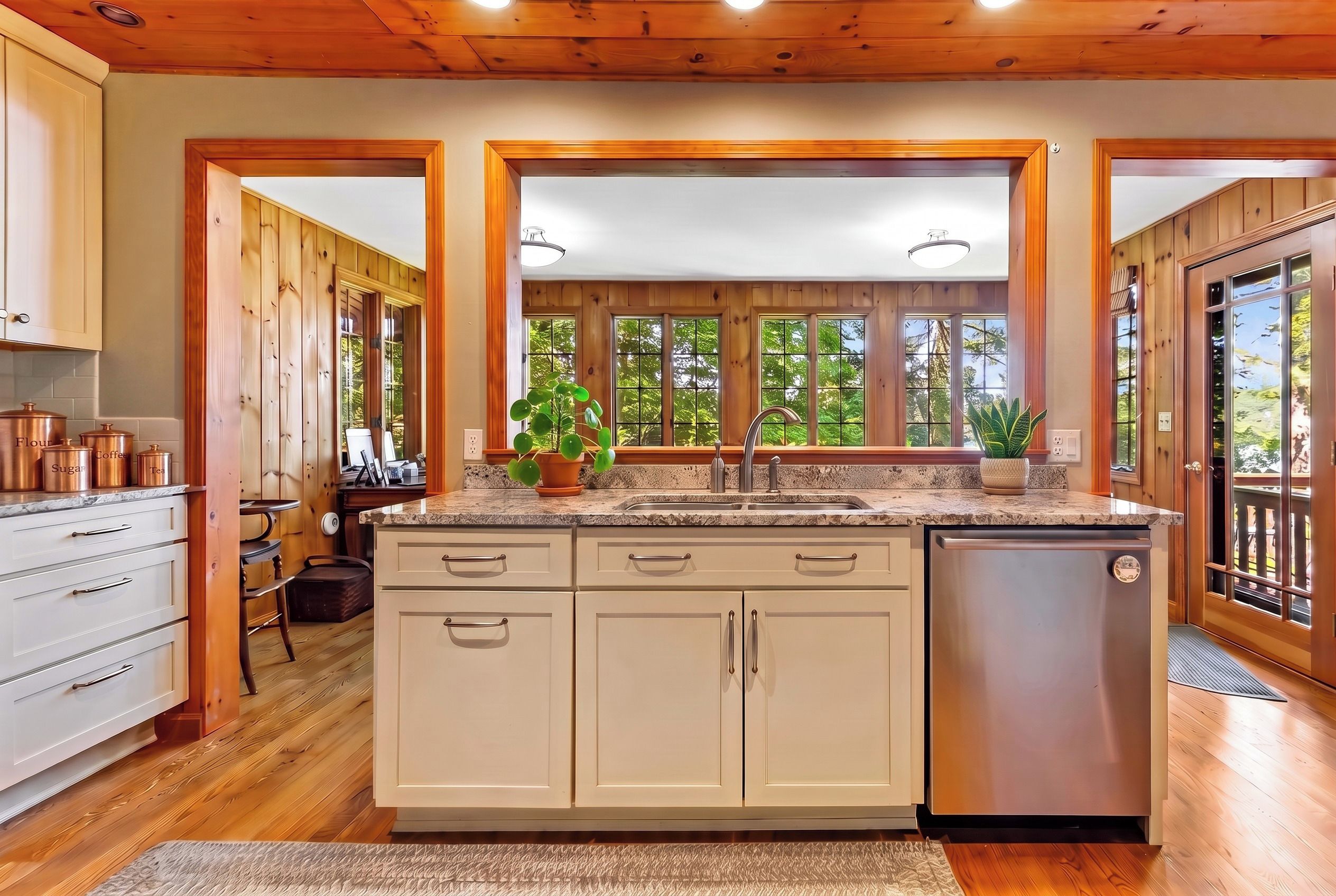 A kitchen with wooden cabinets, a stainless steel dishwasher, and a window with a view of trees.