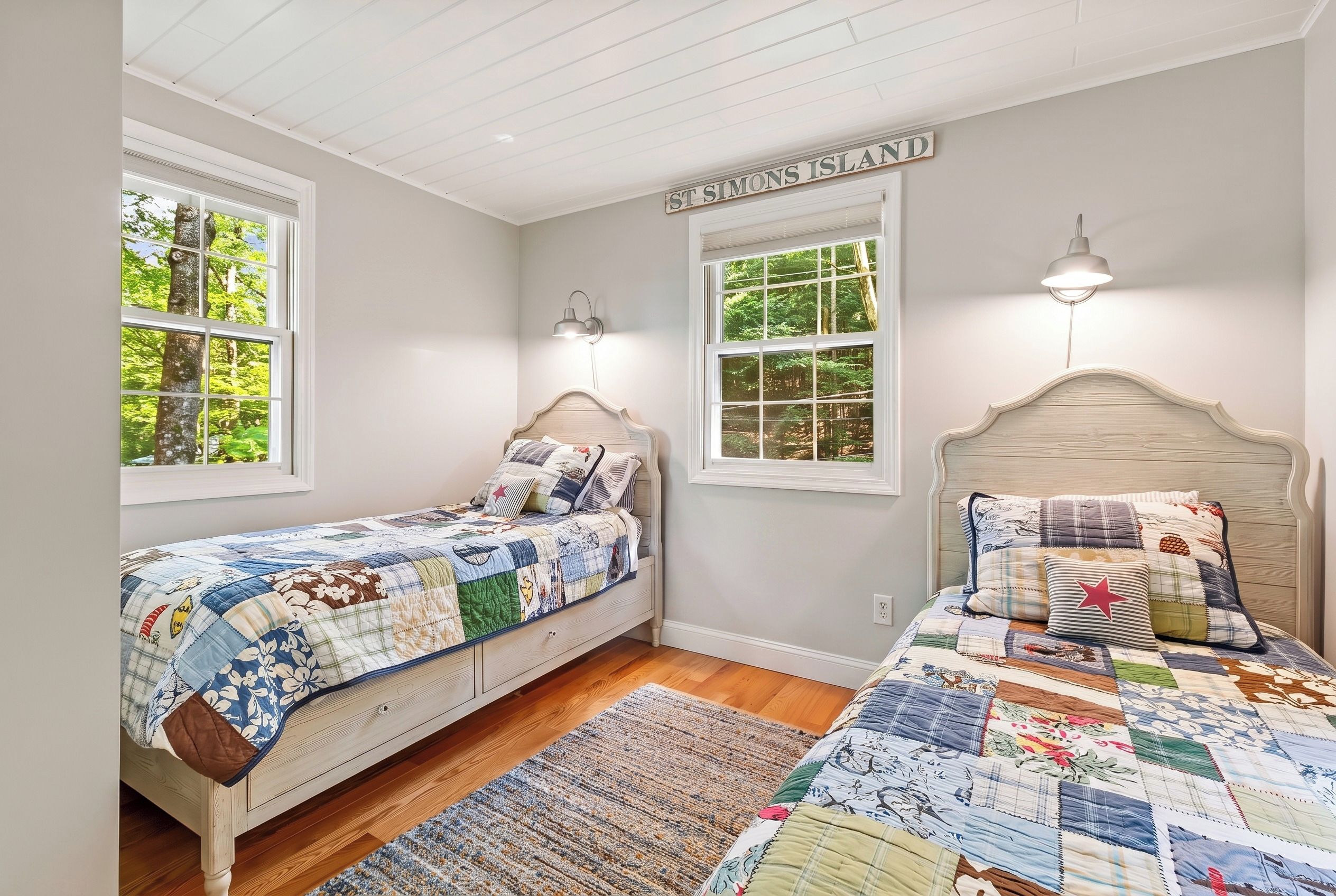 A bedroom with two beds, a wooden headboard, a quilted bedspread, and a view of trees through the windows.