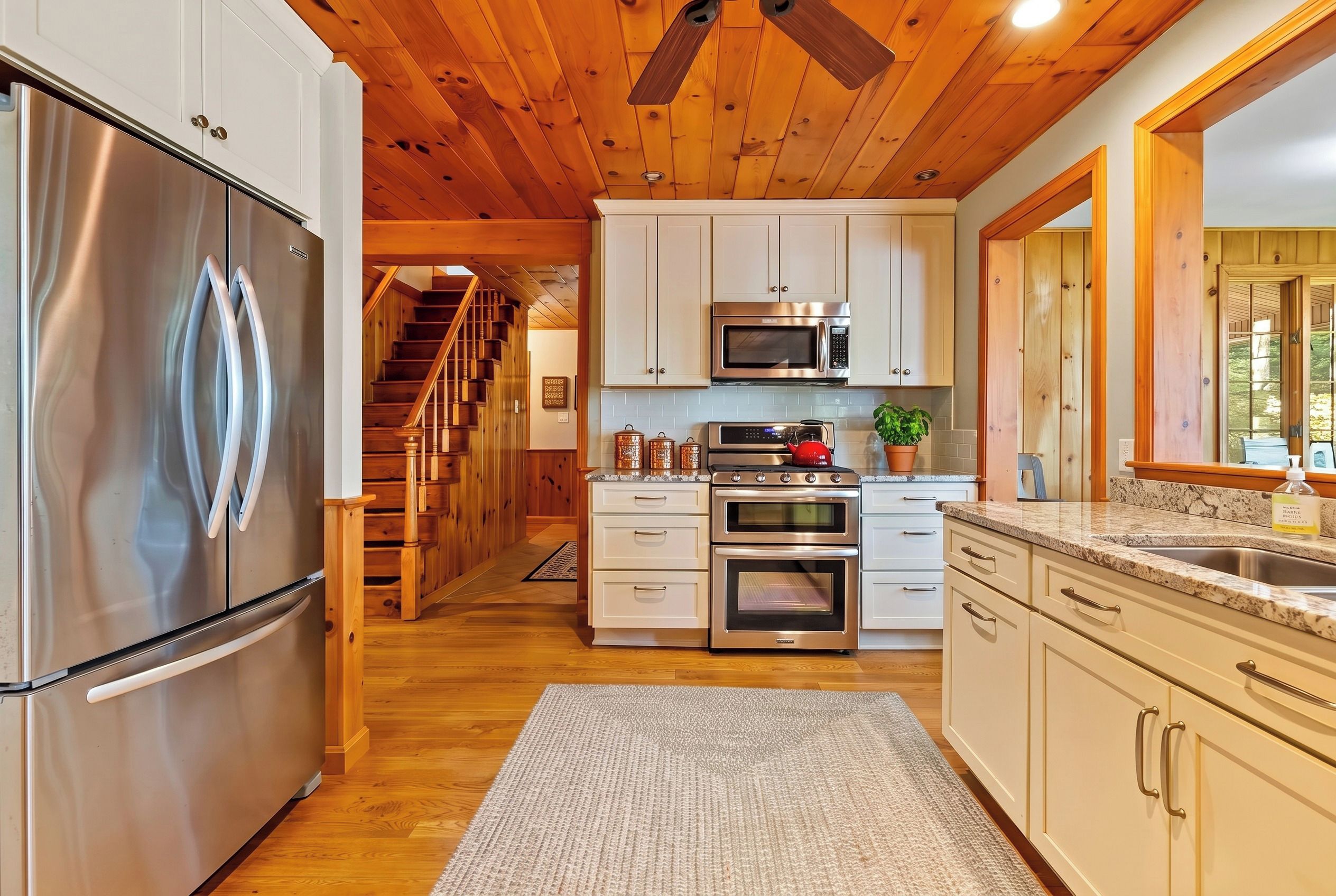 A modern kitchen with stainless steel appliances, wooden flooring, and a view of a wooden staircase leading to another level.