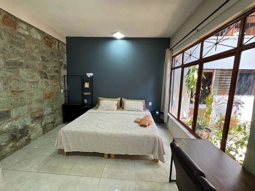Un dormitorio con una cama grande, una mesita de noche, una lámpara y vistas al exterior a través de una ventana.