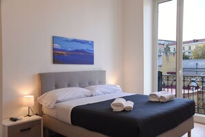 Een hotelkamer met een bed, nachtkastje, lamp, schilderij van een stadsgezicht en uitzicht op gebouwen door een raam.