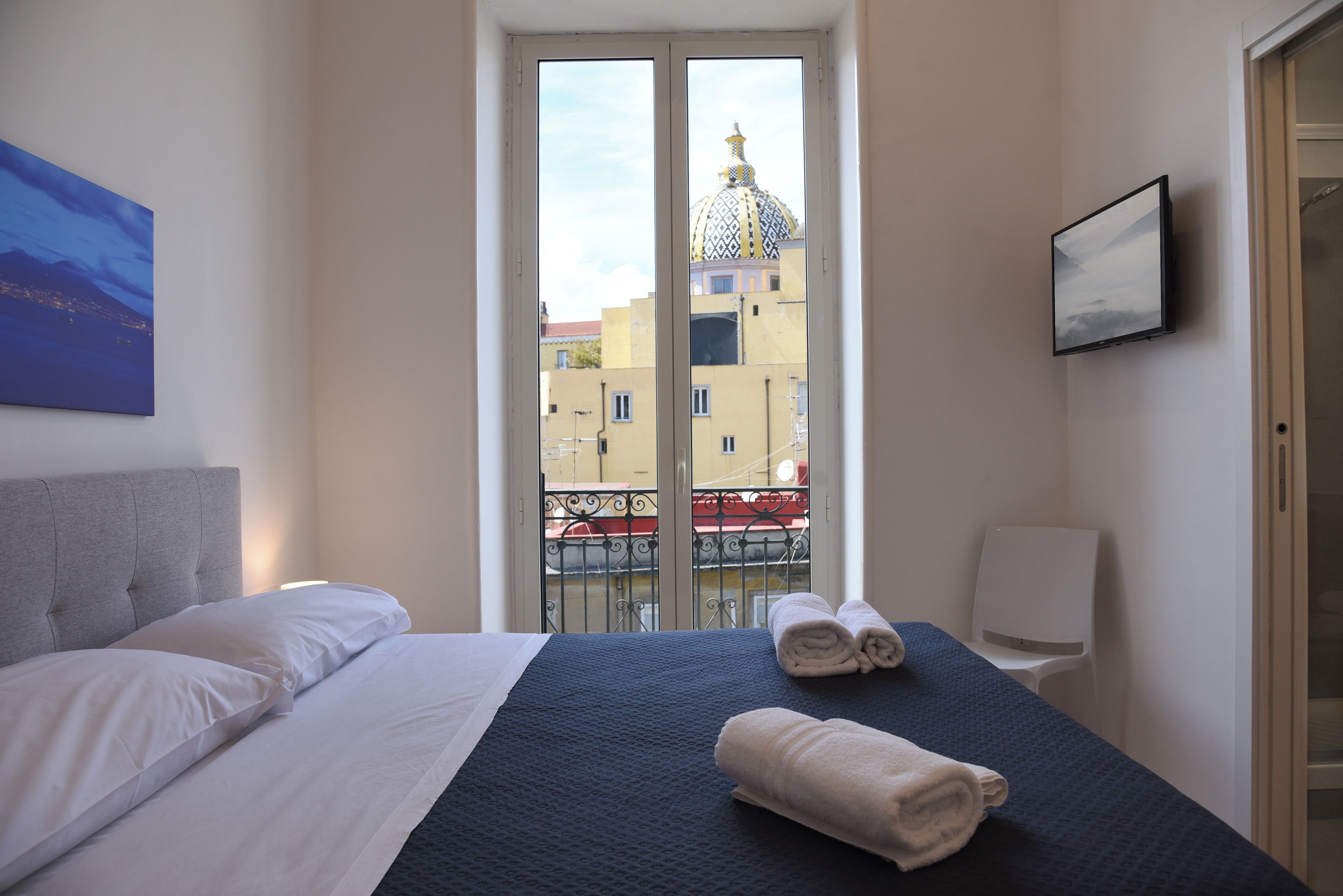 A hotel room with a bed, a TV, a balcony with a view of buildings, and a painting on the wall.