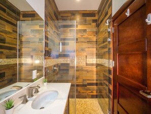 Un baño moderno con ducha envuelta en vidrio, un lavamanos blanco con dos grifos y una puerta de madera.