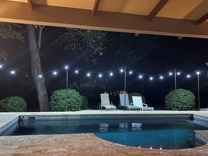 Piscina con solárium empedrado, rodeada de follaje y sillas de descanso blancas.