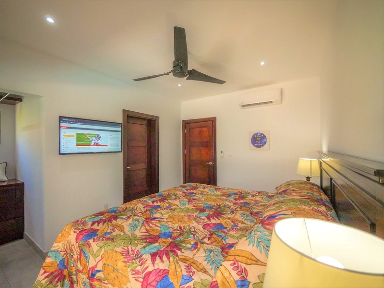 Quarto com cama, ventilador de teto, televisão na parede e uma porta.