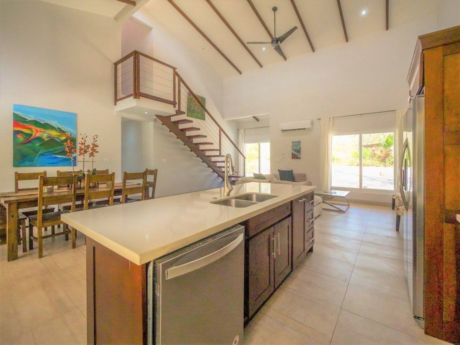 A modern kitchen with a central island, a dining area with wooden chairs, a staircase, and a view of the outdoors.
