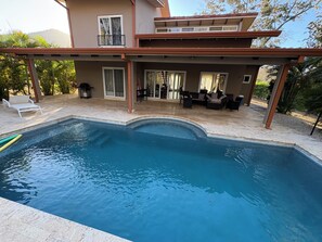 Una piscina con un porche techado, mobiliario exterior y vistas a un edificio con varias ventanas.