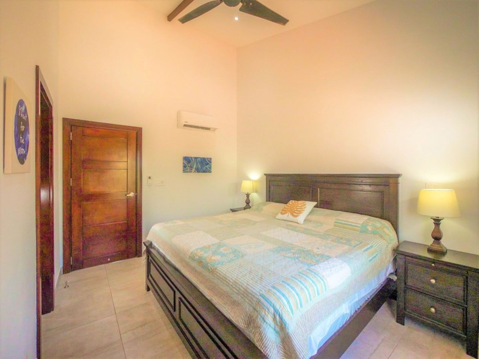 A bedroom with a bed, two bedside tables, a ceiling fan, a door, and a wall-mounted air conditioning unit.