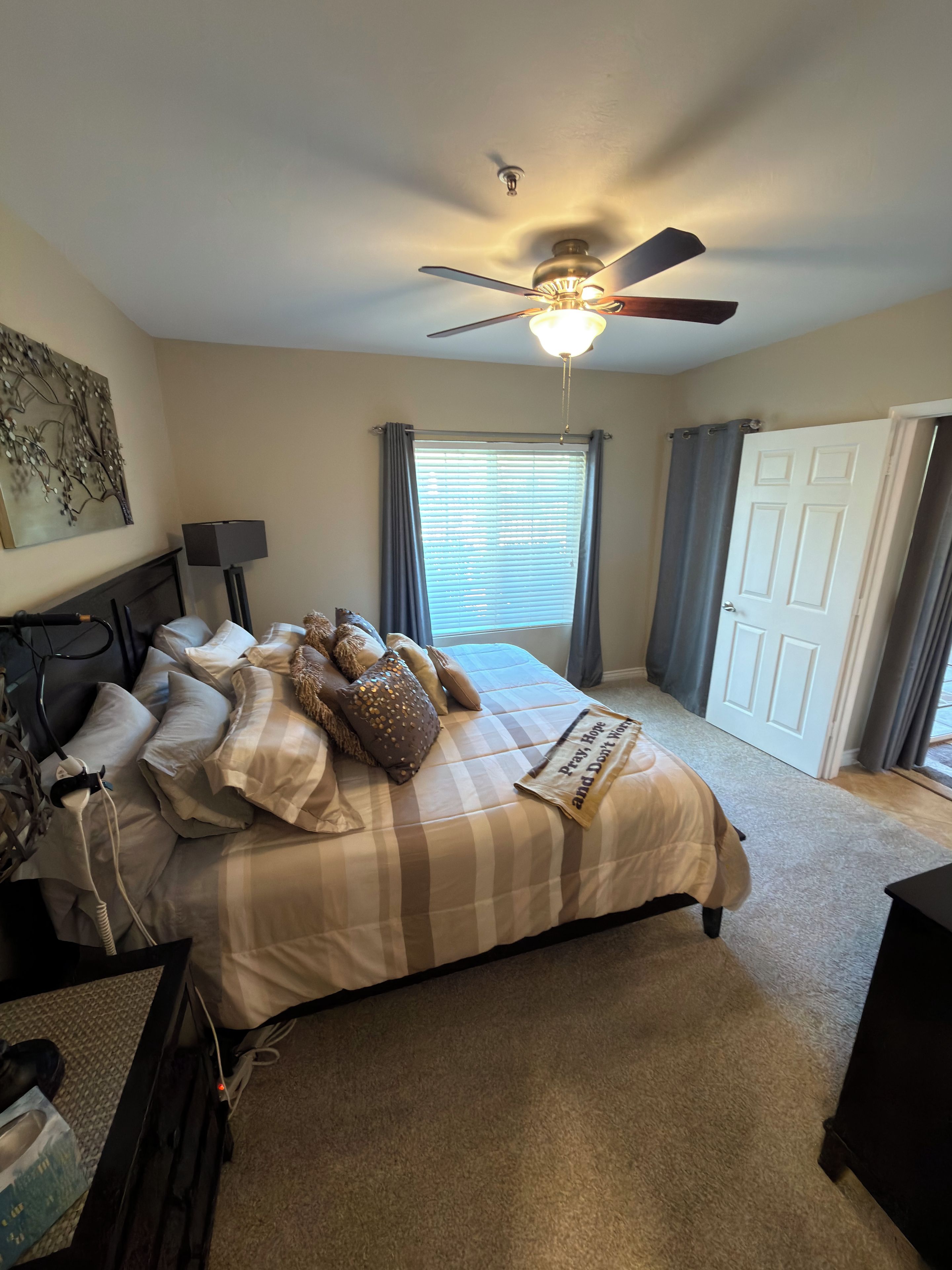 A bedroom with a bed, a ceiling fan, a window with curtains, a door, and a wall-mounted lamp.