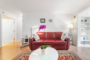 Ein rotes Ledersofa mit gemusterten Kissen, ein weißer Couchtisch mit einem Topf und Pflanze, eine Stehlampe und ein Bücherregal mit Dekoration.