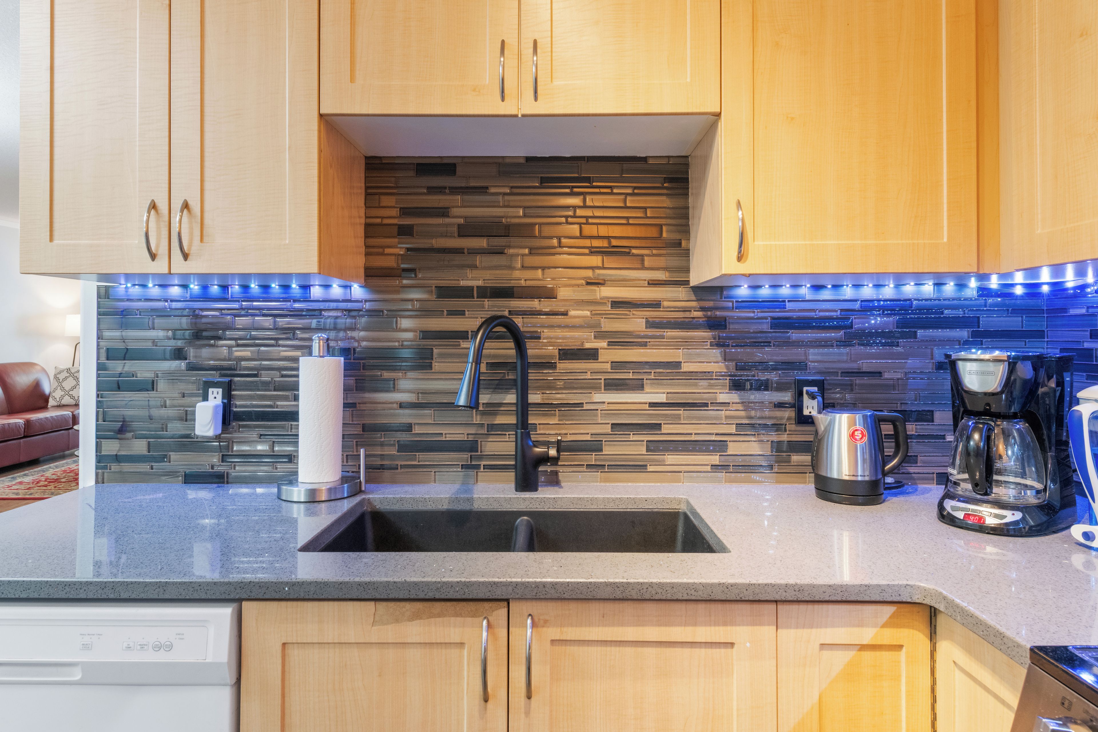Una cocina con armarios de madera, salpicadero de azulejos, un lavamanos de acero inoxidable y una cafetera.
