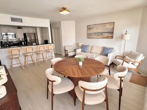 Un salon moderne avec une table ronde en bois, six chaises, un canapé et une cuisine en arrière-plan.