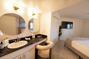 Une chambre d’hôtel dotée d’un grand miroir rond, d’un lavabo avec des robinets dorés, d’une table de chevet, d’un lit et d’un téléviseur fixé au mur.