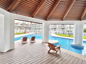 Un espace piscine abrité, doté d’une terrasse en bois, de chaises longues et offrant une vue sur un bâtiment du complexe hôtelier.