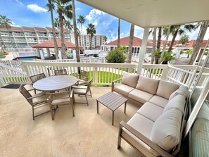 A covered outdoor seating area with a round table and chairs, a sofa, and a view of a hotel building and palm trees. - 6 Beds | Pool Steps Away | Private Beach Access (South Padre Island)